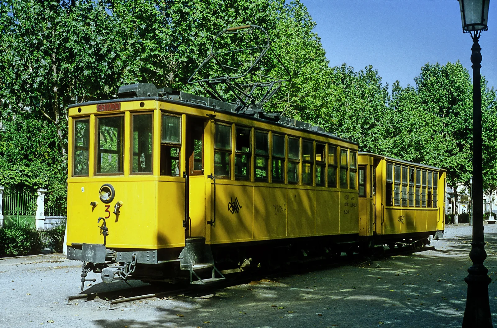 Coche motor amarillo número 3 del tranvía de Sierra Nevada, con su pantógrafo y un remolque detrás, expuesto bajo los árboles junto a una farola en Granada