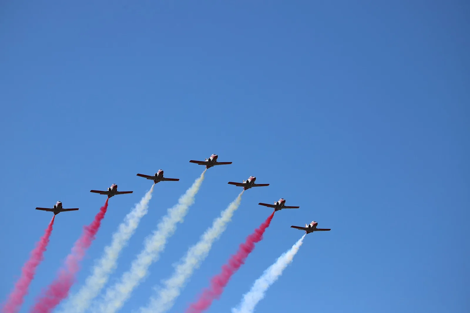Siete aviones de la Patrulla Águila en formación sobre un cielo azul, dejando estelas de humo rojo y amarillo durante la Fiesta Nacional de 2020 en Madrid.