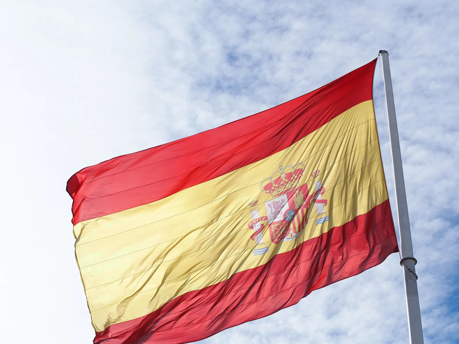 Bandera de España de gran tamaño ondeando en su mástil contra un cielo azul con nubes, en la plaza de Colón de Madrid.