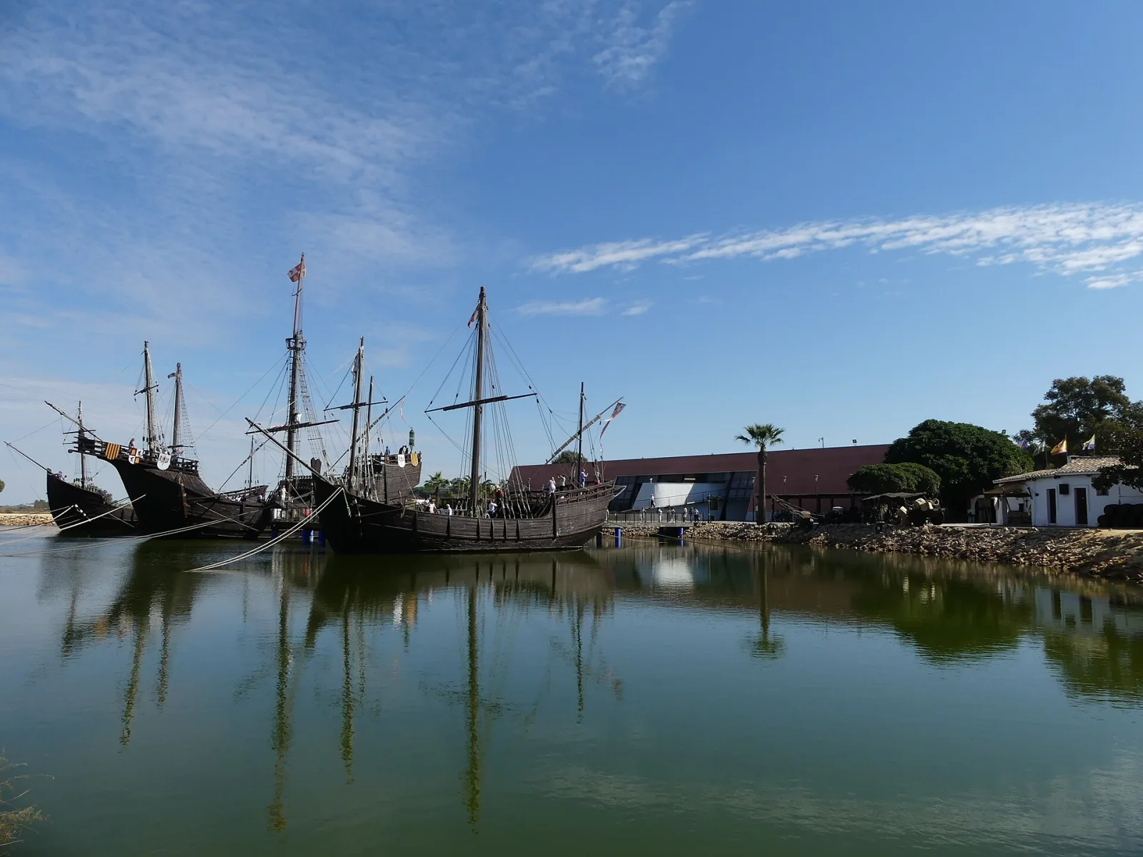 Las tres réplicas de las naves de Colón, fondeadas al fondo de una lámina de agua quieta que las refleja, bajo un cielo azul con nubes.