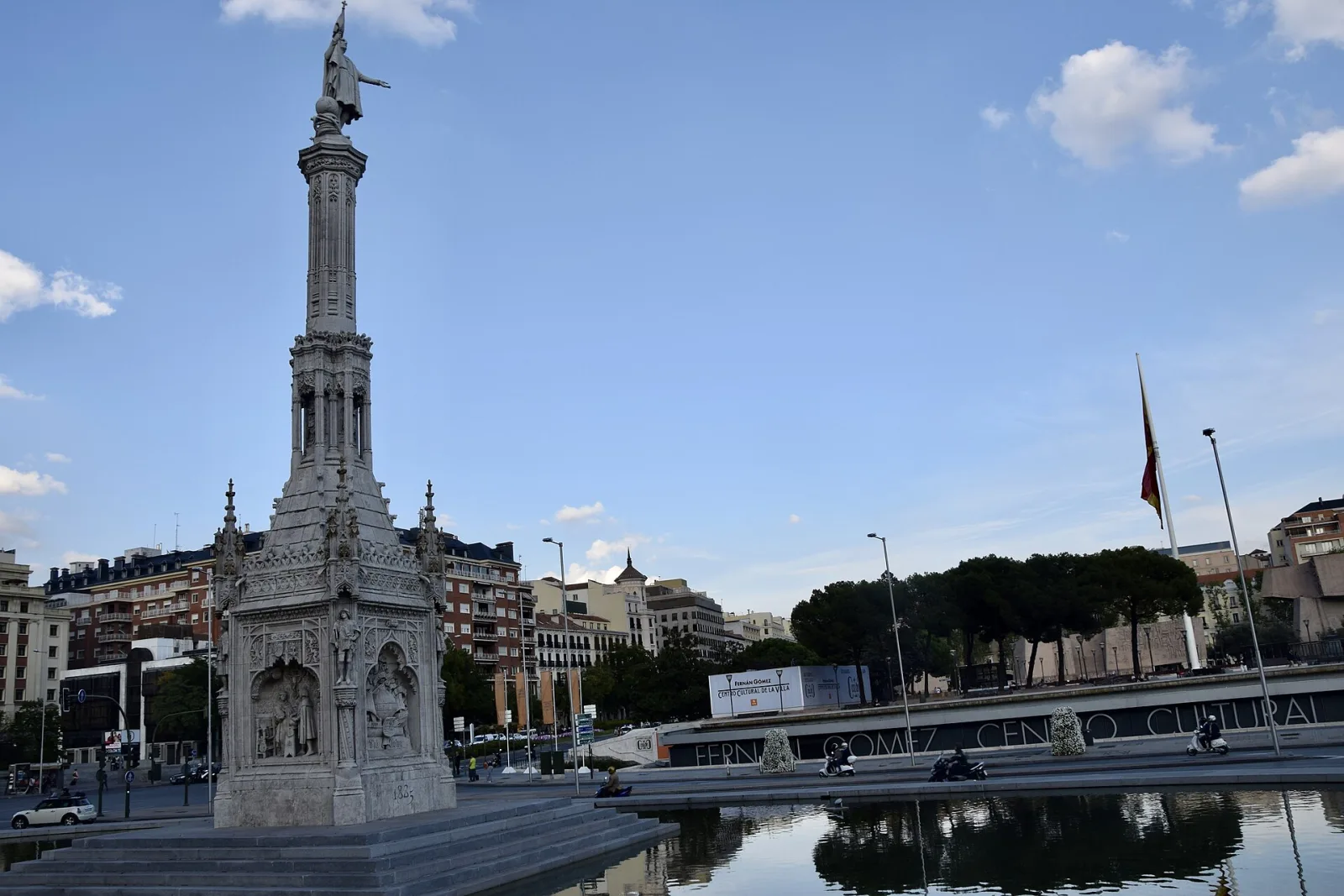 Columna neogótica coronada por la estatua de Colón en la plaza de Colón de Madrid, con una lámina de agua y edificios al fondo bajo un cielo azul.