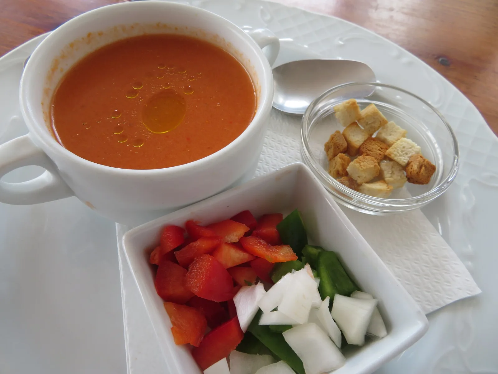 Taza de gazpacho junto a un cuenco con tomate, pimiento y cebolla picados y otro con picatostes, sobre un plato blanco.