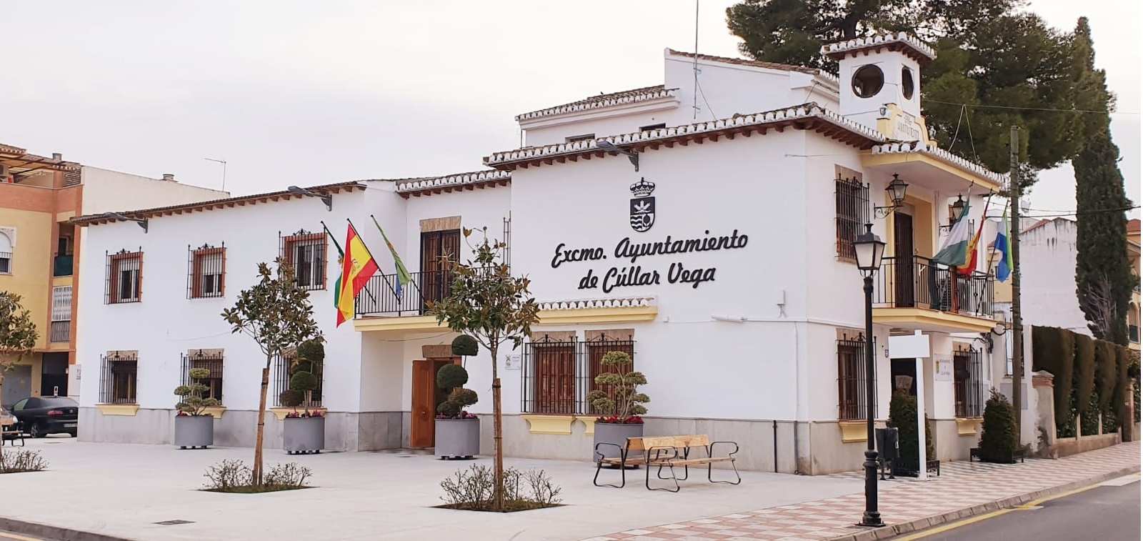 Edificio blanco de dos plantas con el rótulo Excmo. Ayuntamiento de Cúllar Vega, banderas en balcones y una plaza con bancos y árboles en macetas.