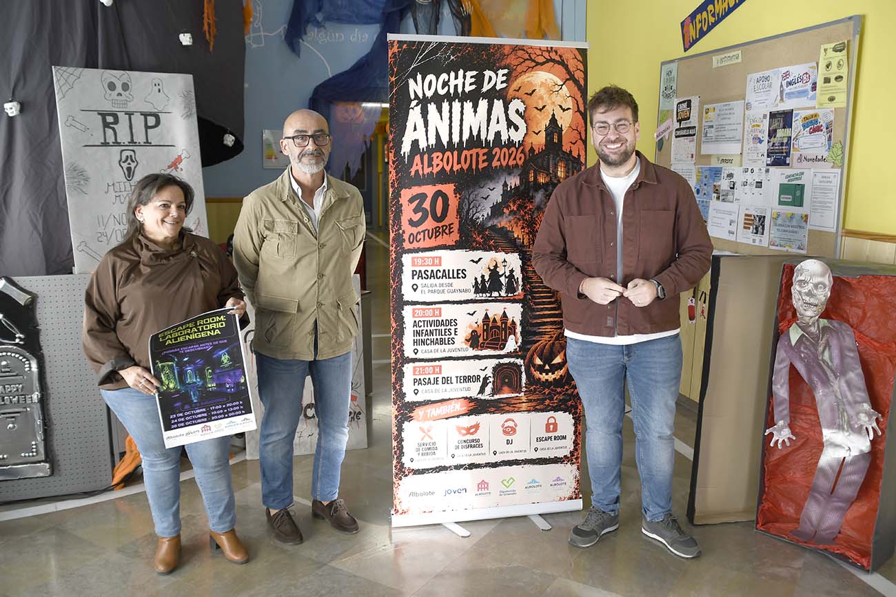 Tres personas de pie junto a un cartel vertical de la Noche de Ánimas y decoraciones de Halloween en una sala.