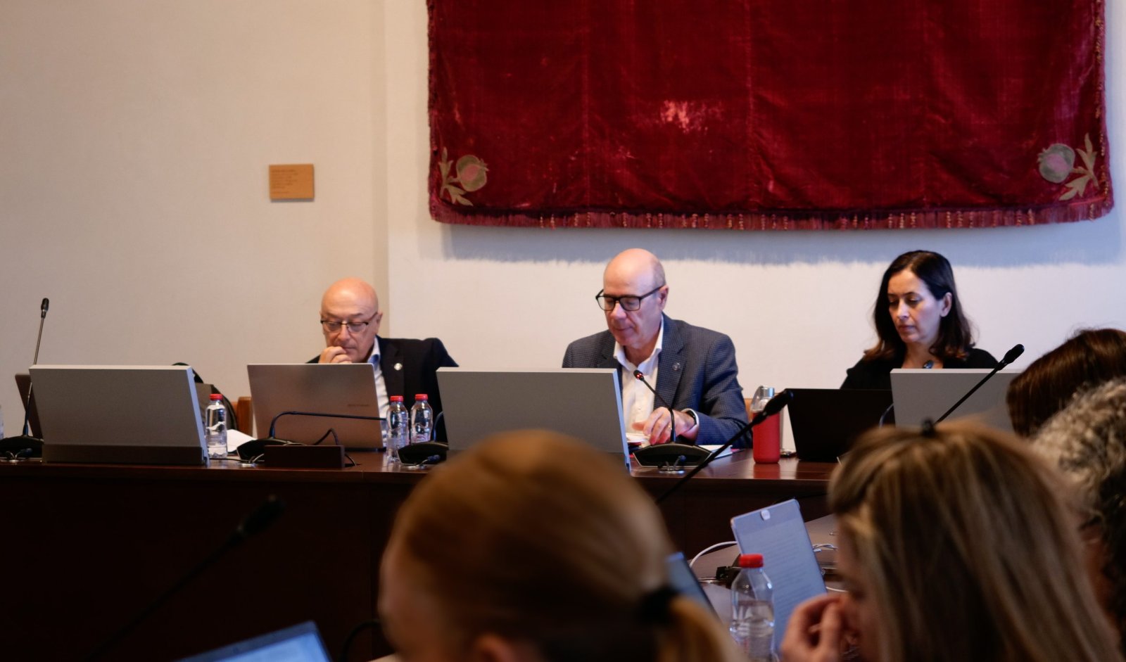 Tres personas con portátiles sentadas tras una mesa en una sala de reuniones, con un tapiz rojo al fondo y público en primer plano.