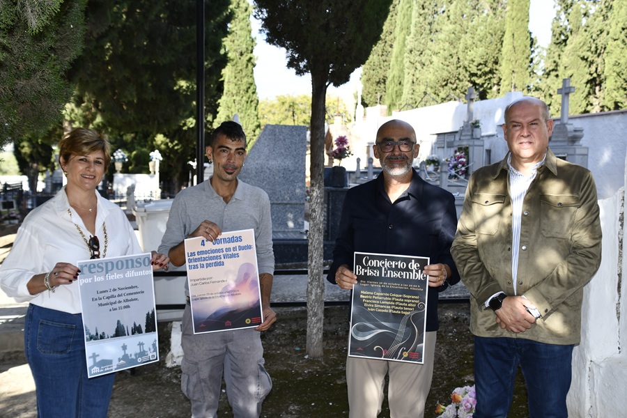 Cuatro personas de pie en un cementerio sostienen carteles con textos sobre actos conmemorativos, entre tumbas y cipreses.
