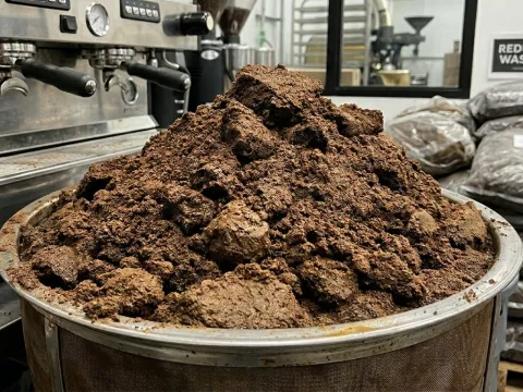 Un gran montón de posos de café húmedos en un recipiente metálico, junto a una cafetera industrial y sacos al fondo.