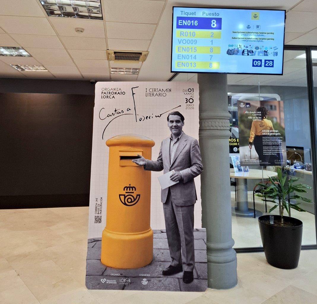 Un cartel recortable con un hombre trajeado junto a un buzón amarillo, en un vestíbulo con pantalla de turnos.