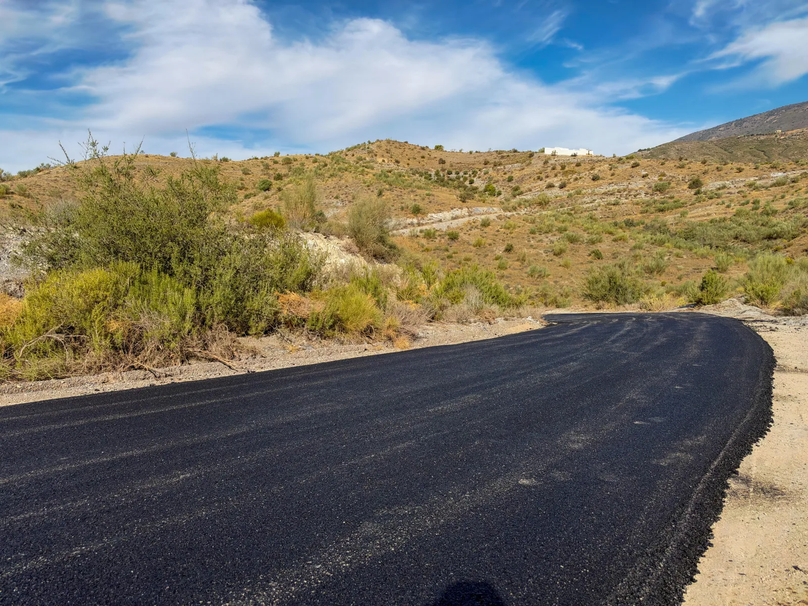 Un camino de asfalto negro recién puesto que curva entre matorrales secos, con colinas áridas y una construcción blanca al fondo bajo un cielo azul con nubes.