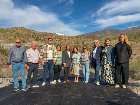 Diez personas posan de pie sobre un camino de asfalto negro recién extendido, con colinas secas y un cielo azul con nubes al fondo.