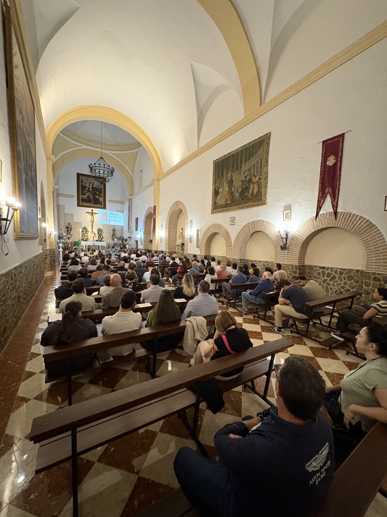 Numerosos asistentes sentados en bancos de madera dentro de una nave eclesiástica con bóvedas, arcos y cuadros.
