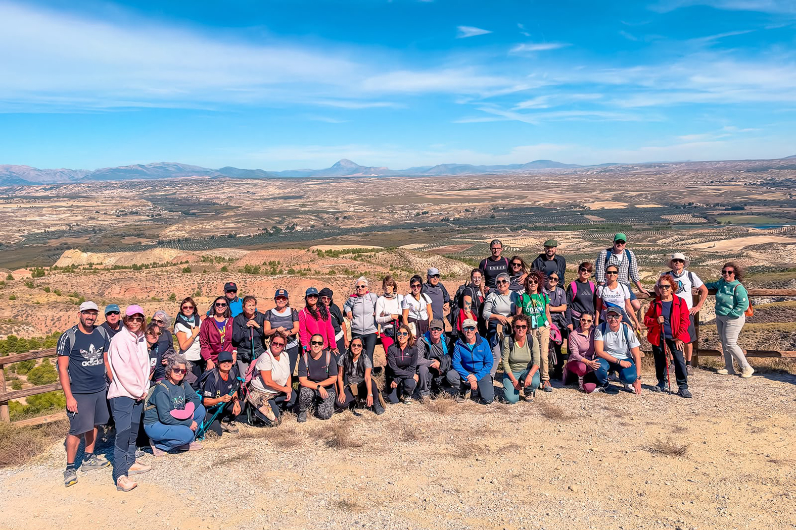 Un grupo de unas cuarenta personas posa en un mirador de tierra junto a una valla de madera, con un amplio valle de campos y montañas al fondo bajo cielo azul.