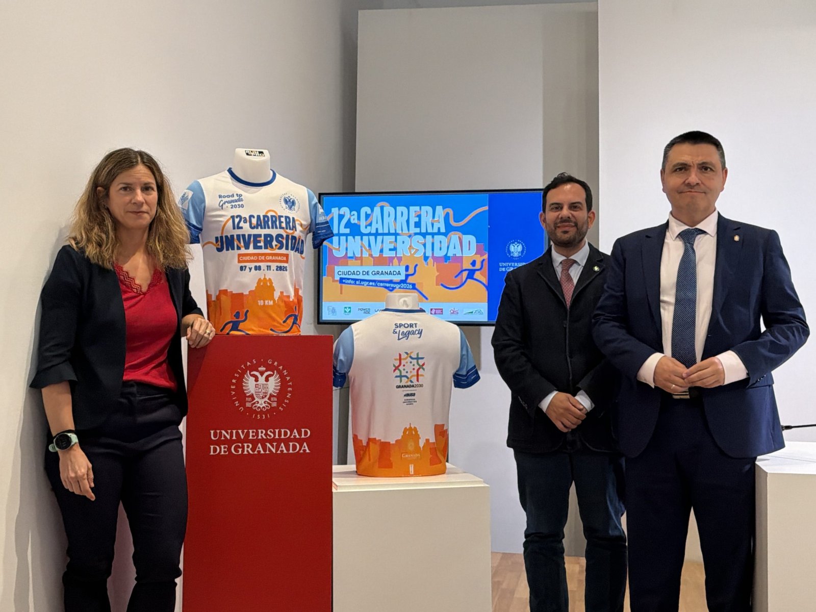 Tres personas de pie junto a dos camisetas deportivas expuestas y una pantalla con el texto 12ª Carrera Universidad.