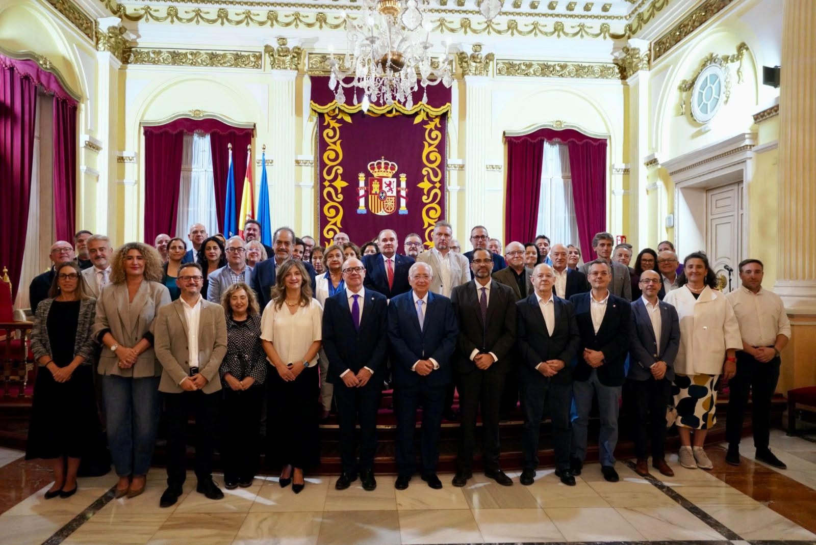 Un grupo numeroso de personas trajeadas posa de pie en una sala noble con cortinajes rojos, banderas y un escudo en la pared.
