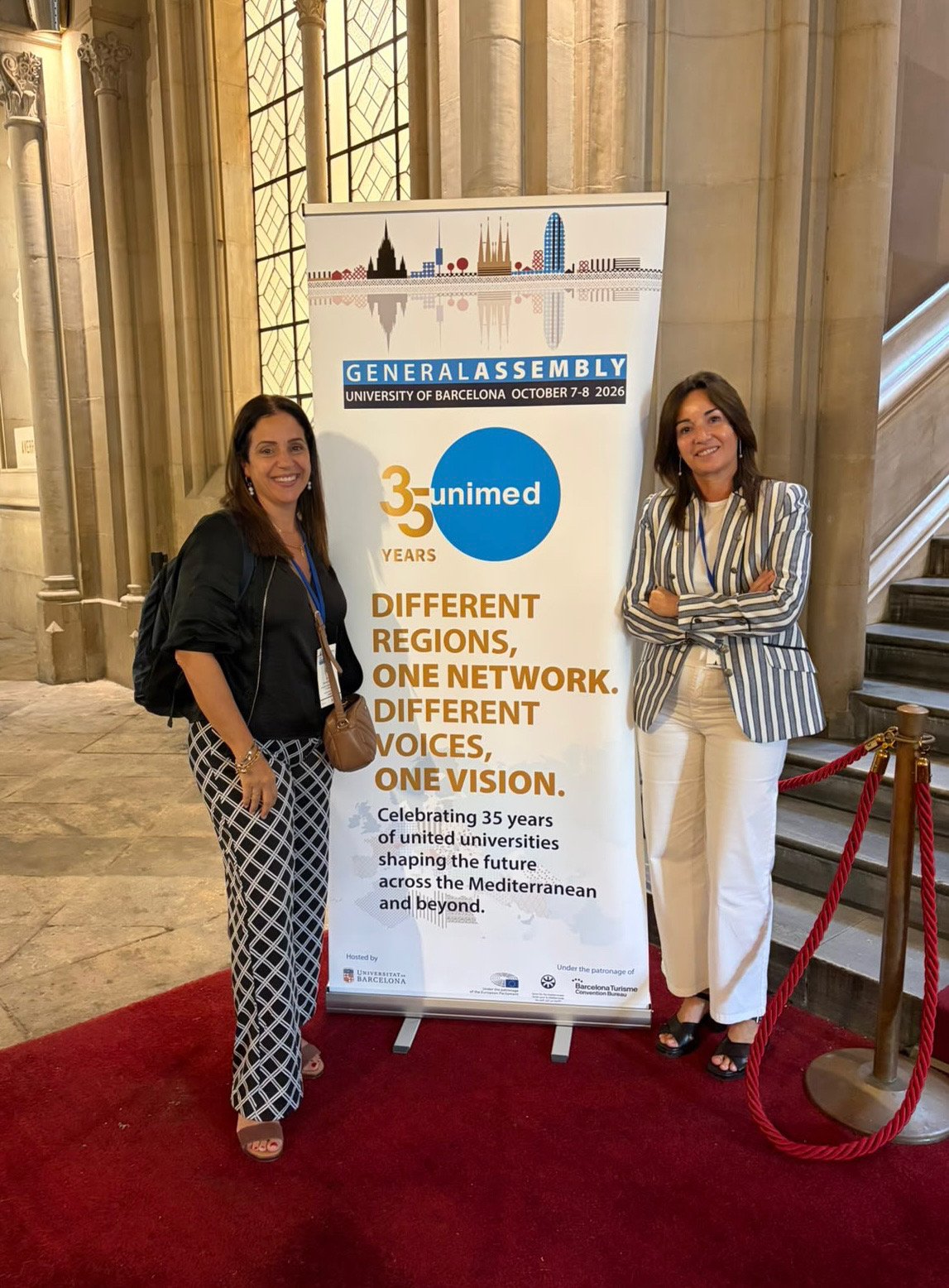 Dos mujeres de pie junto a un cartel de unimed en un interior con columnas de piedra y alfombra roja.
