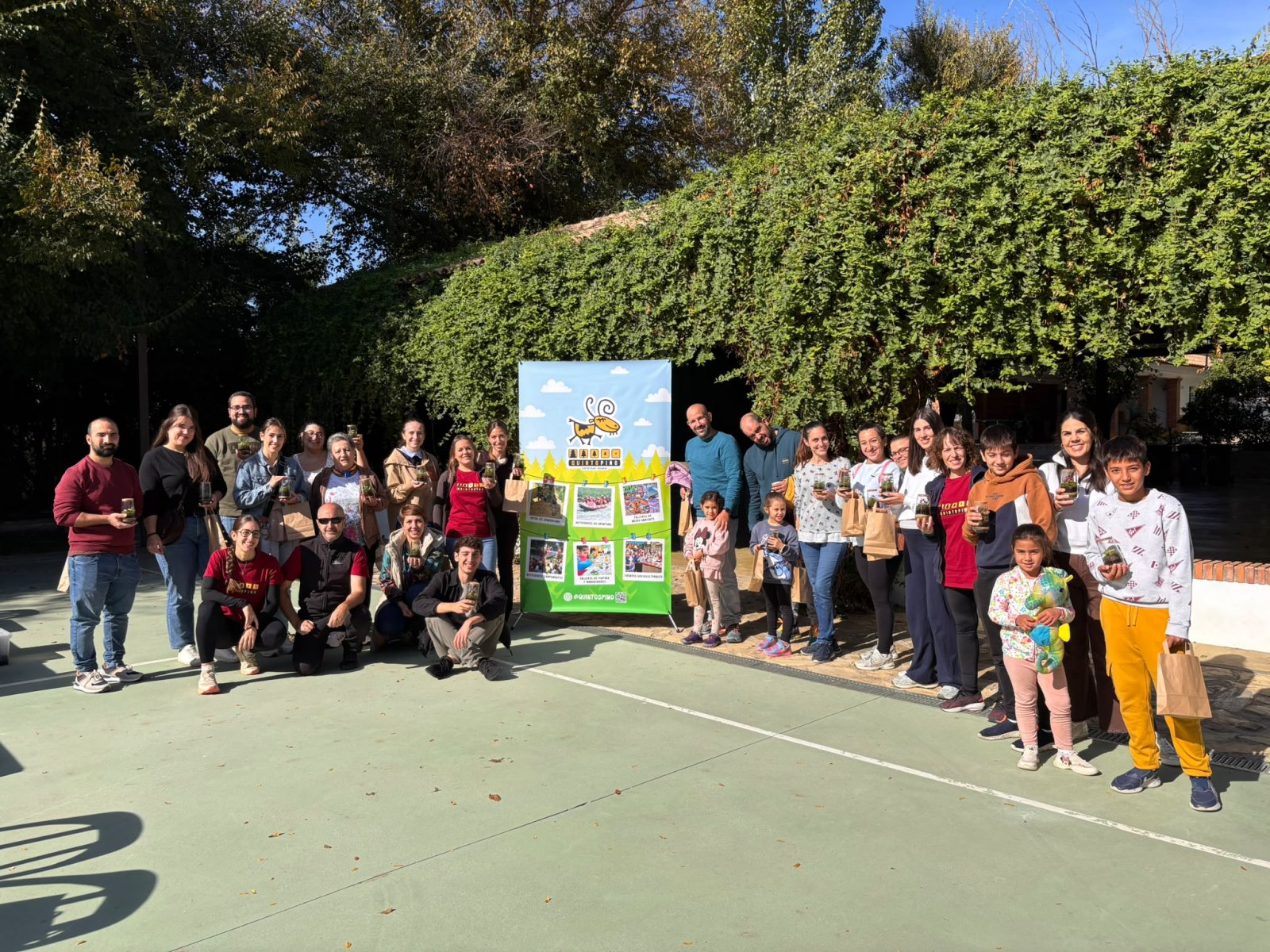 Un grupo de personas de distintas edades posa al aire libre en una pista deportiva, sosteniendo pequeños tarros y junto a un cartel verde.