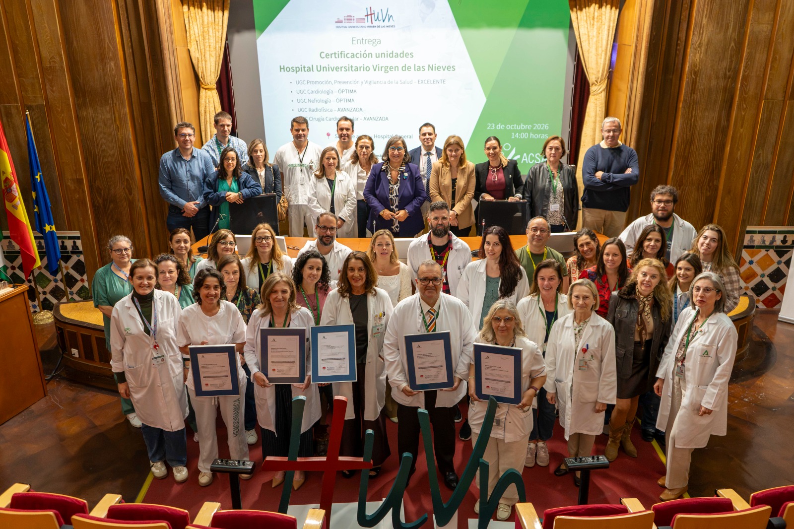 Un grupo de personas con batas blancas y trajes posa en un salón con banderas y una pantalla que indica la entrega de certificaciones de calidad.