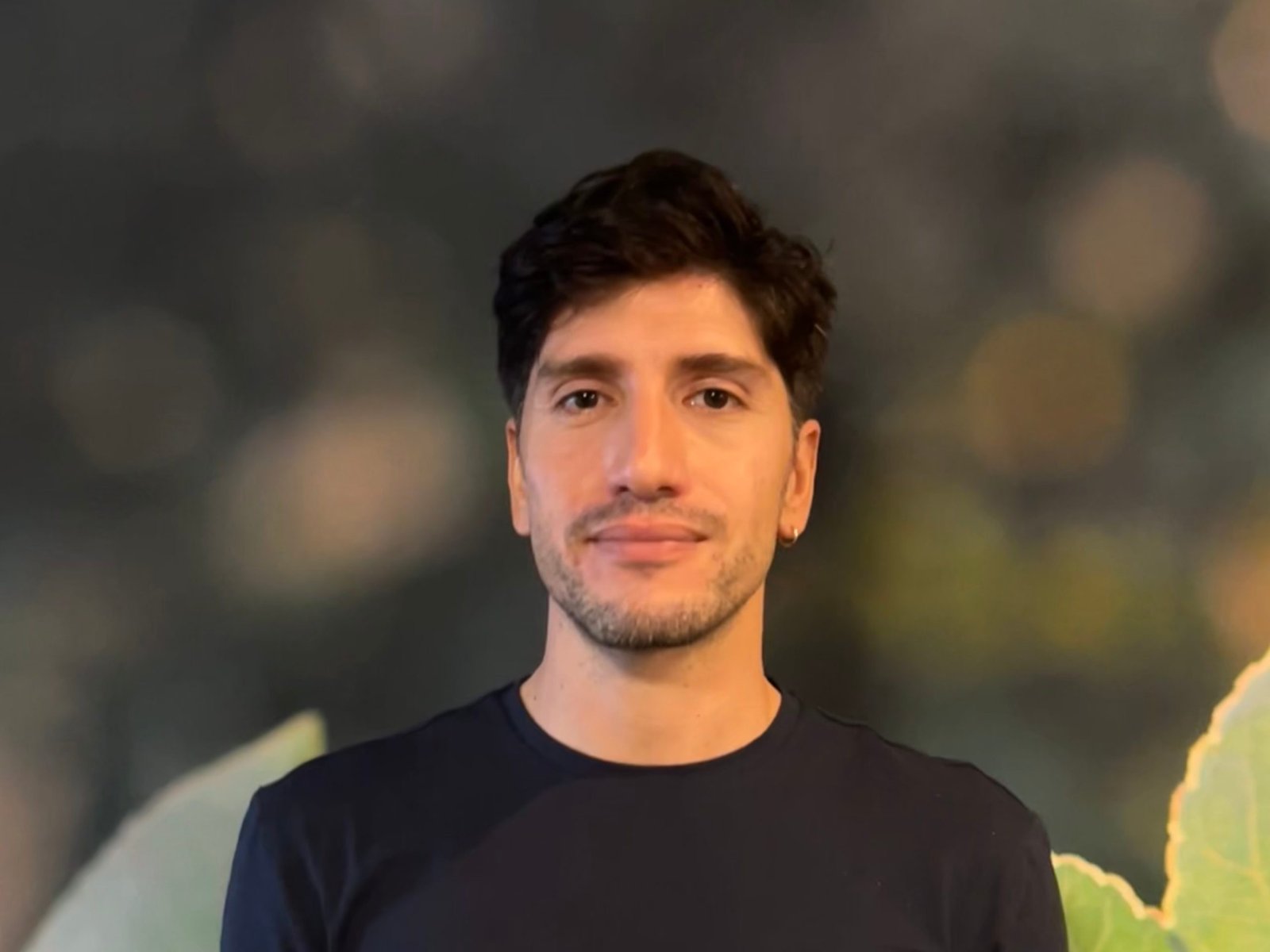 Primer plano de un hombre joven de pelo oscuro y barba incipiente, con camiseta azul marino, sobre un fondo desenfocado.