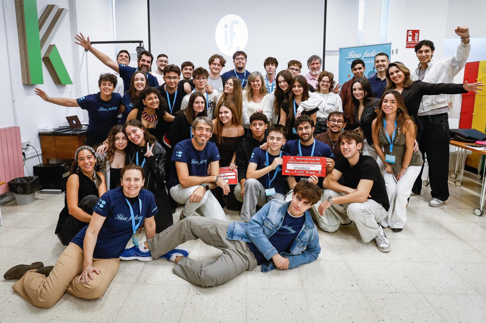 Un grupo numeroso de jóvenes posa sonriente en una sala, algunos con camisetas azules y acreditaciones, y uno sostiene un diploma rojo.