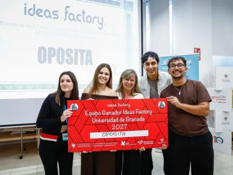 Cinco personas jóvenes posan sonrientes sosteniendo un gran cartón rojo con el texto Equipo Ganador Ideas Factory Universidad de Granada 2027 OPOSITA, ante una