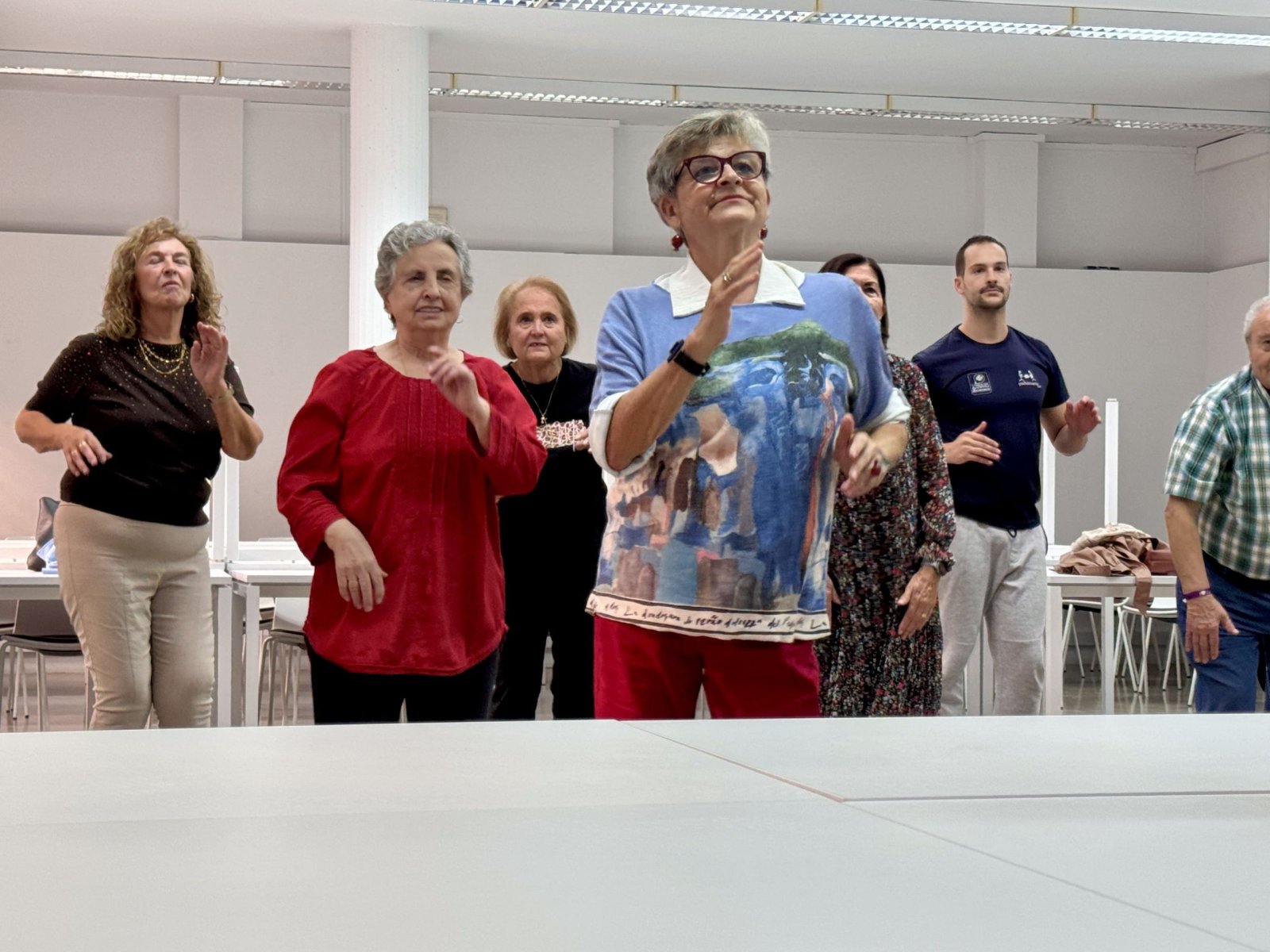 Un grupo de personas mayores y un joven, de pie en una sala blanca, con los brazos en movimiento como si bailaran.
