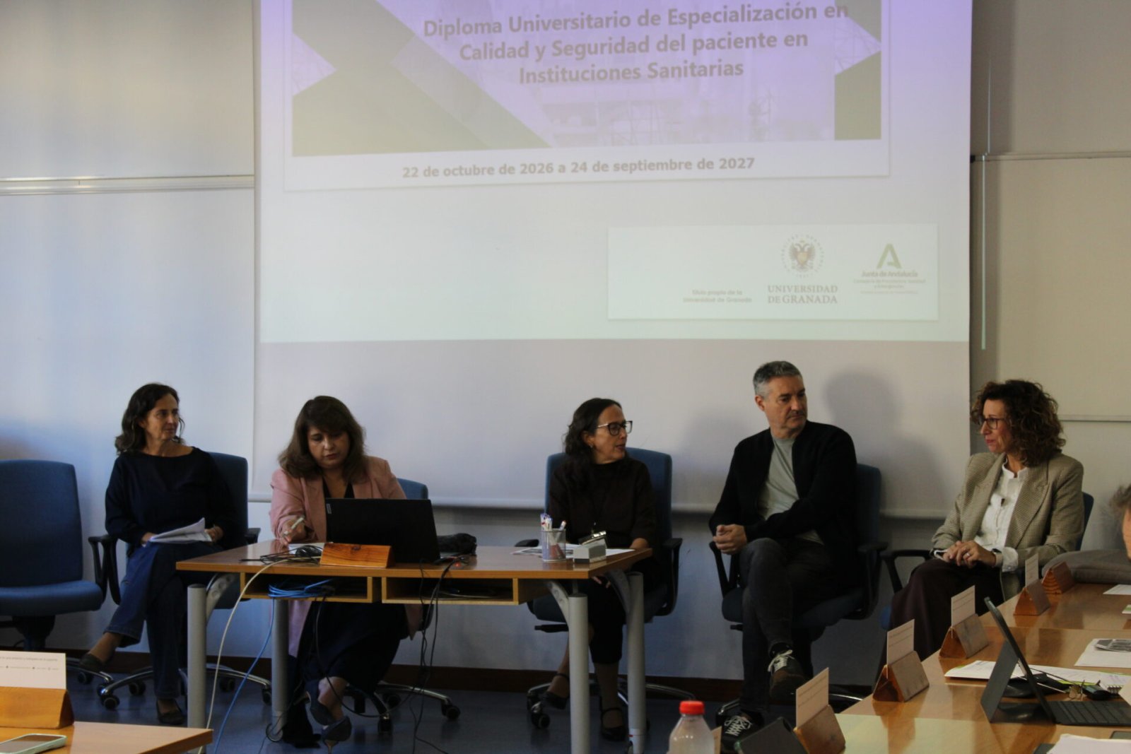 Cinco personas sentadas tras una mesa en una sala, con una proyección sobre un diploma universitario de la UGR al fondo.