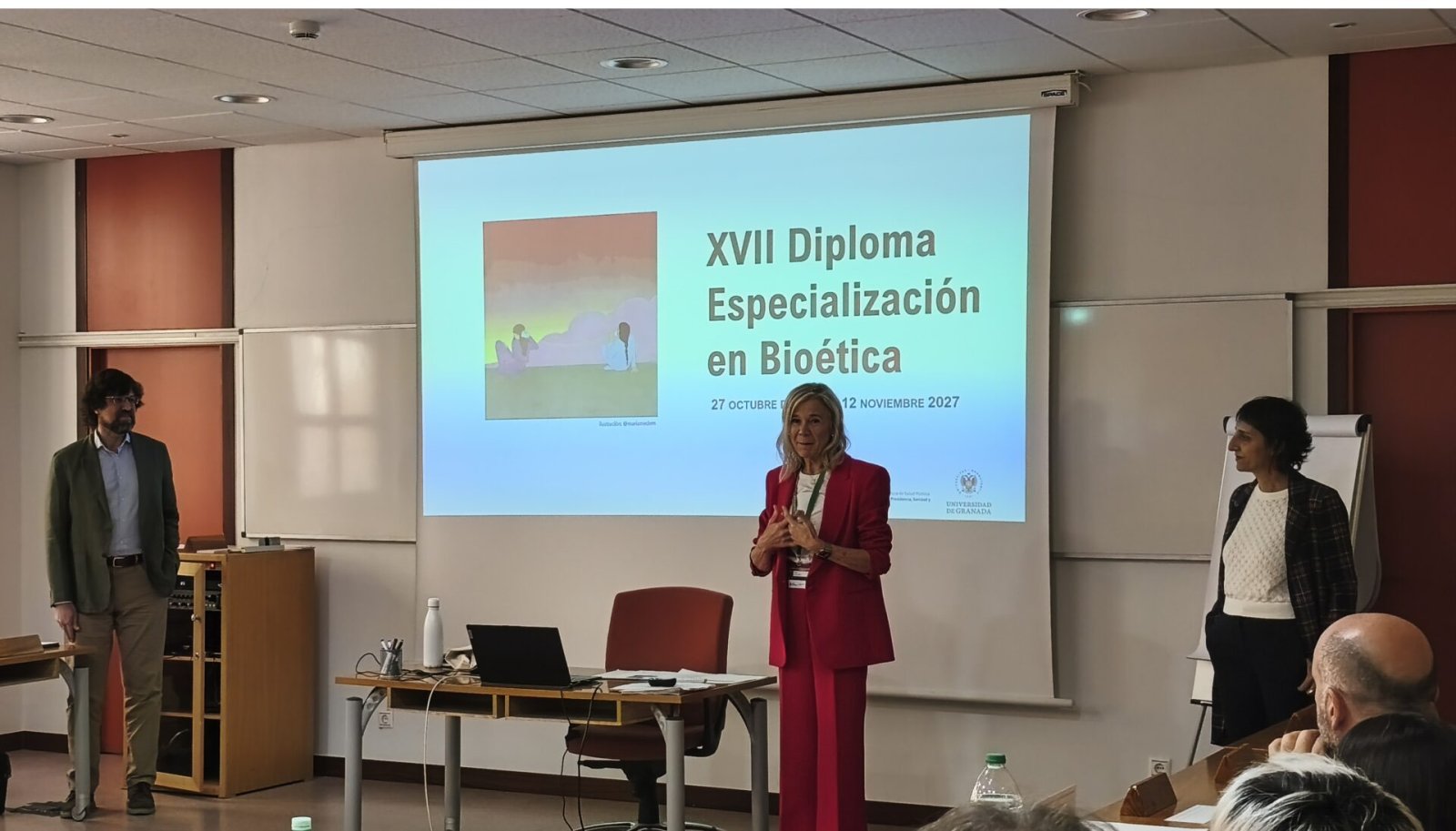 Una mujer con traje rojo habla ante una pantalla que proyecta XVII Diploma Especialización en Bioética, con dos personas más de pie en un aula.