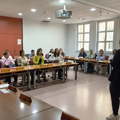 Un grupo de personas sentadas en mesas de un aula escucha a alguien de pie que habla junto a una ventana.