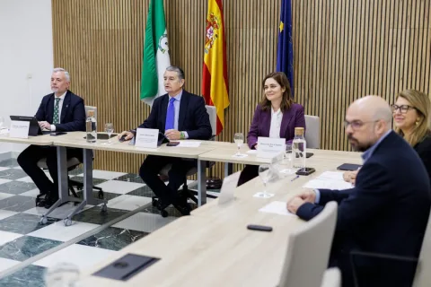 Cinco personas trajeadas sentadas en una mesa de reuniones, con banderas de Andalucía, España y la UE al fondo.