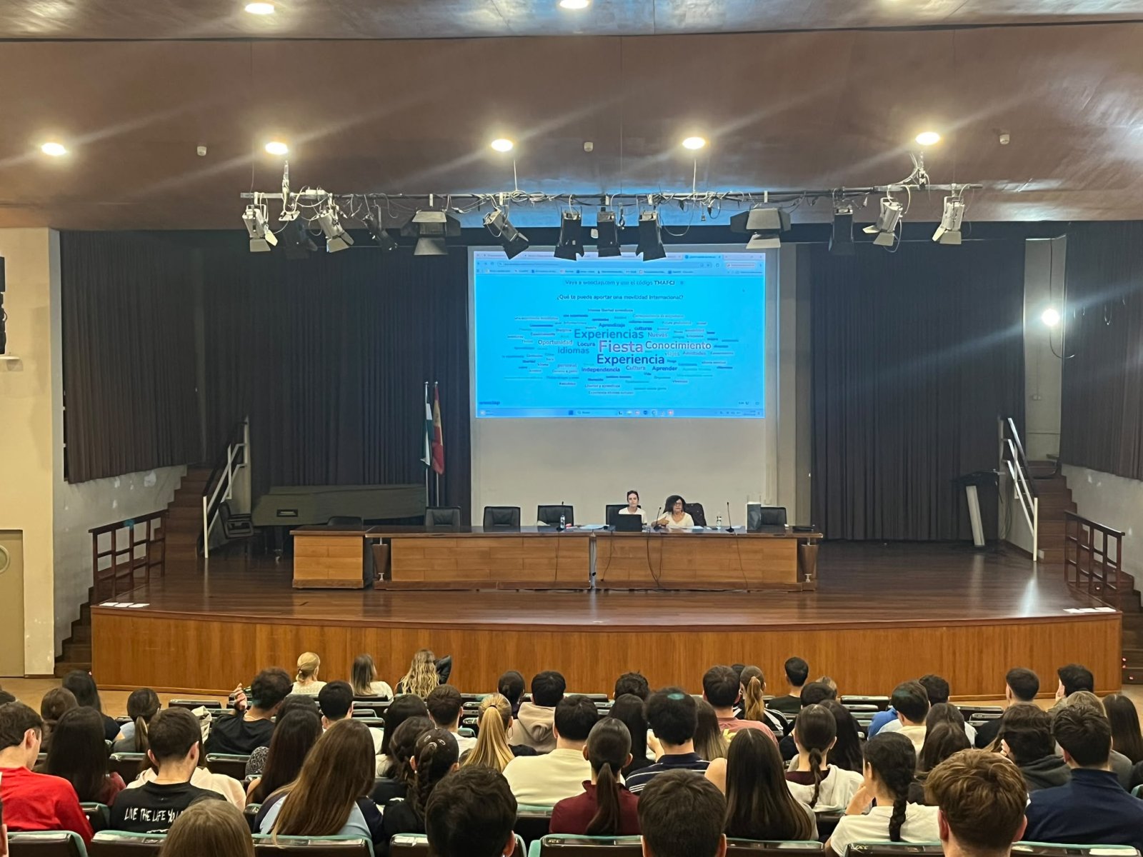 Un auditorio con público sentado frente a un escenario donde dos personas hablan tras una mesa y una pantalla proyecta texto.