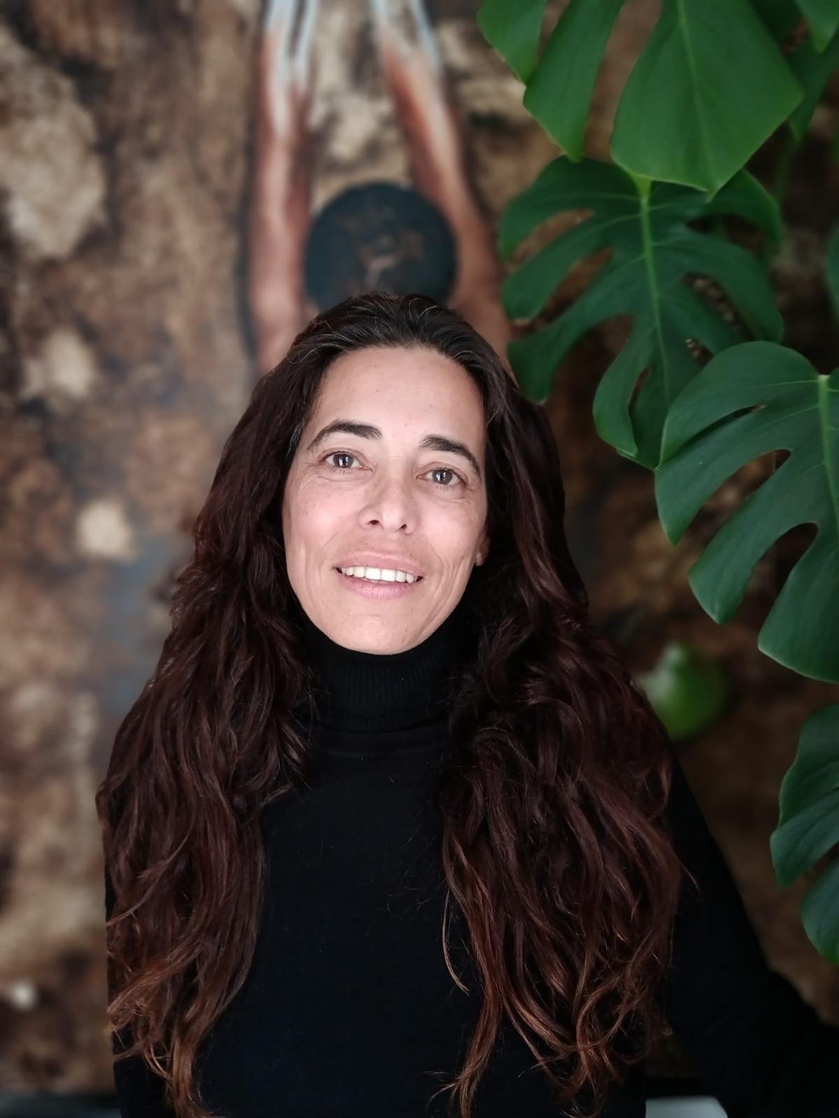 Mujer de pelo largo y oscuro con jersey negro de cuello alto, sonriendo, junto a una planta de hojas grandes y un fondo desenfocado.