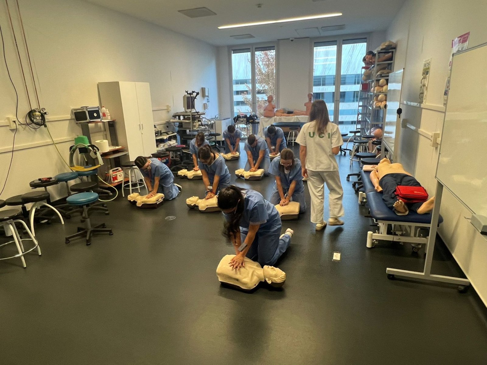 Varias personas con pijama sanitario practican reanimación sobre maniquíes en el suelo de una sala amplia con camillas y equipo médico.