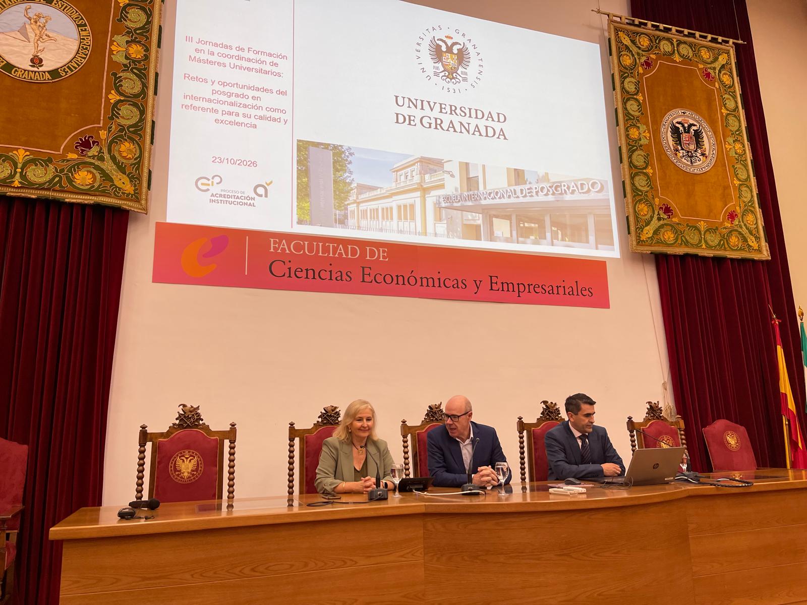 Tres personas sentadas tras una mesa en un salón con pantalla que muestra la Universidad de Granada y un cartel de la Facultad de Ciencias Económicas.