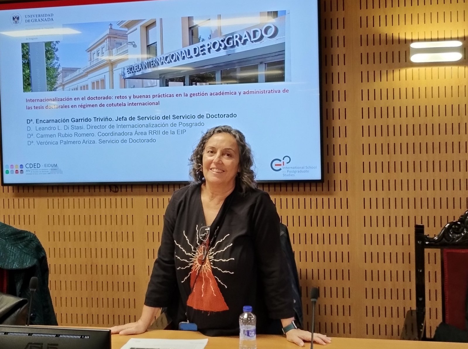 Una mujer de pelo rizado y camiseta negra estampada sonríe tras una mesa, ante una pantalla con una presentación sobre cotutela internacional.