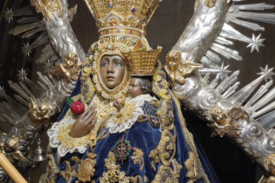 Virgen de rostro oscuro con corona dorada y manto azul bordado, sosteniendo al Niño, sobre un resplandor de rayos plateados.
