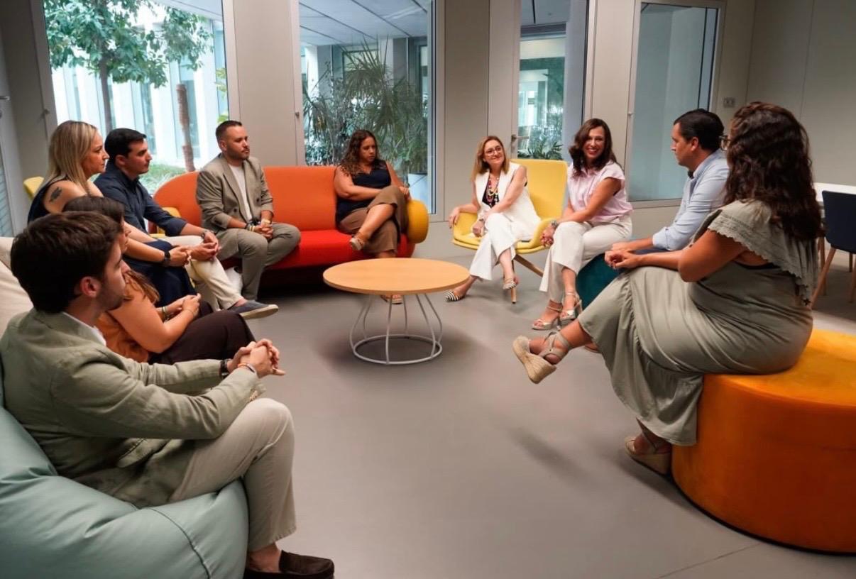 Un grupo de personas sentadas en círculo conversando en una sala luminosa con sofás y pufs de colores.