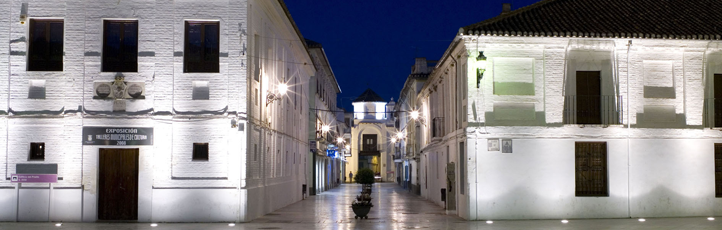 Calle estrecha y vacía de noche entre edificios blancos iluminados, con farolas y un edificio con arco al fondo.
