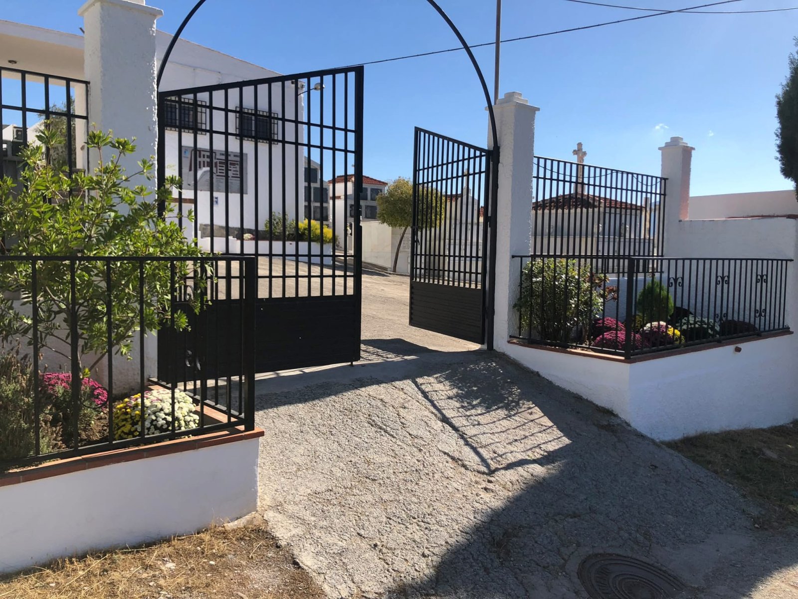 Cancela metálica abierta entre pilares blancos con jardineras, sobre un camino de grava, con edificios y una cruz al fondo.