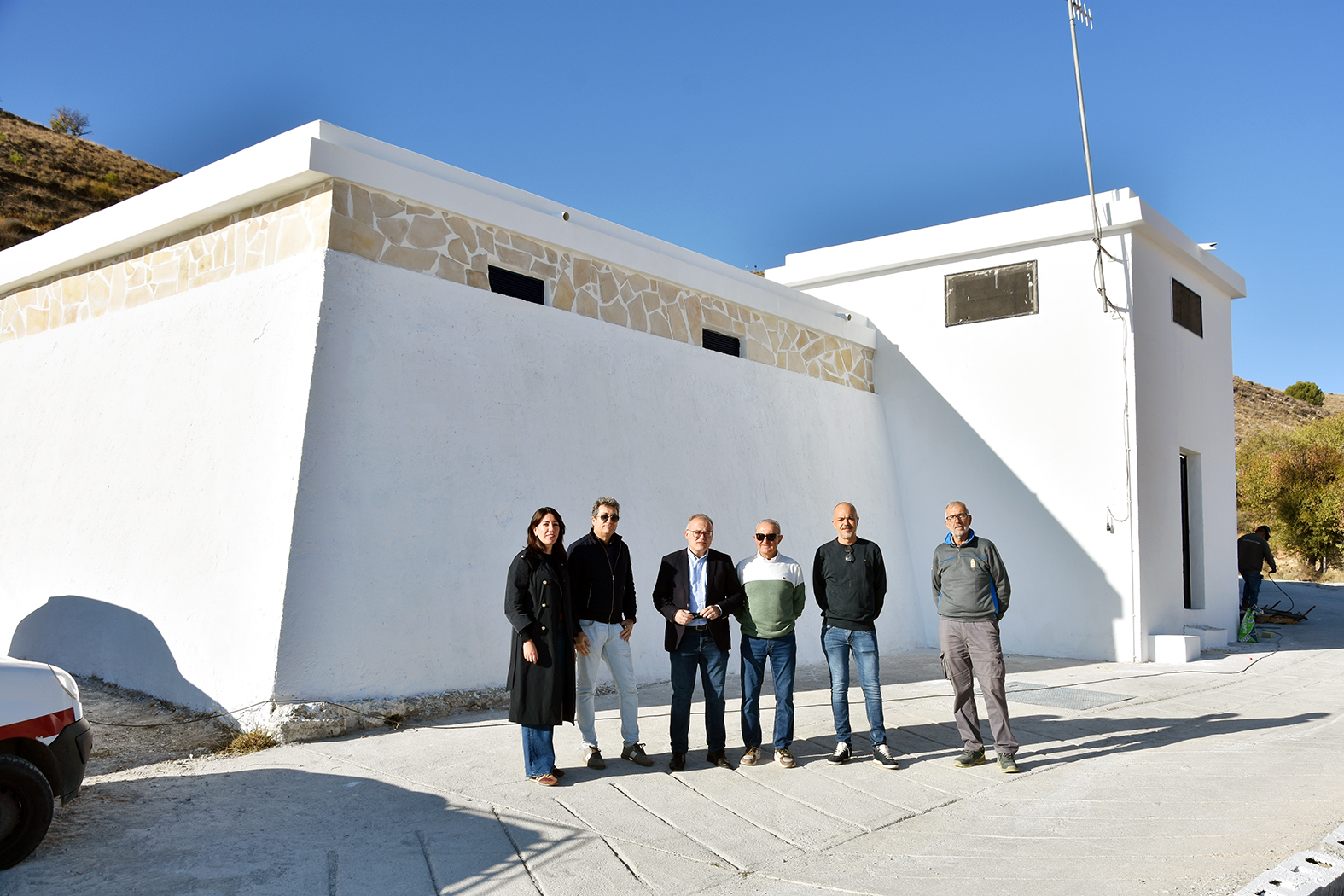 Seis personas de pie junto a un edificio blanco de paredes lisas y remate de piedra, con cielo despejado y una ladera al fondo.